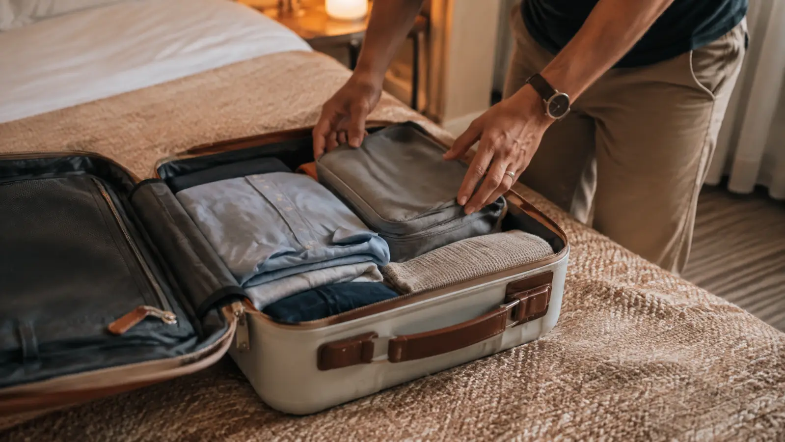 An open suitcase on a bedspread with a traveler's hands rearranging folded clothes and a zippered packing cube before closing the lid