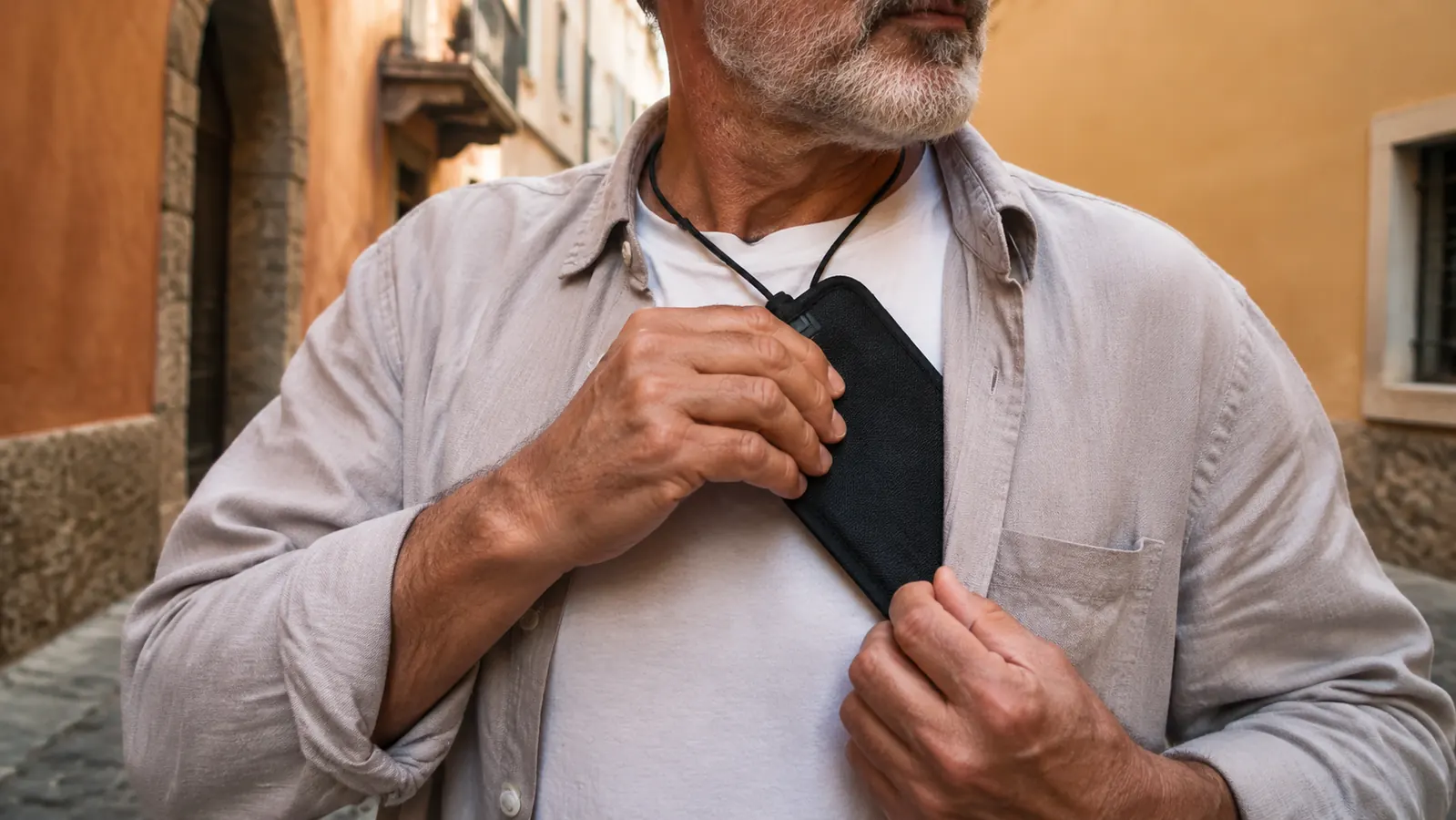 Close-up of a hand tucking the edge of a thin passport wallet flat against a shirt under the collar, a lanyard visible at the neckline, on a bright street.
